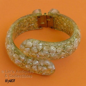 May include: A vintage, clear, hinged bangle bracelet with a gold-colored clasp. The bracelet is filled with gold glitter and small, white, shell-shaped embellishments. The bracelet is on an orange background. The text "Lin's Antiques & Collectibles #1401" is visible.