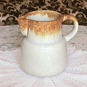May include: A white ceramic pitcher with a brown glaze on the top. The pitcher has a handle and a spout.
