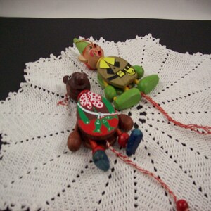 May include: Two wooden pull string toys, one is a brown bear wearing a red and white dress and the other is a green elf wearing a yellow and green outfit. The toys are on a white crocheted doily.