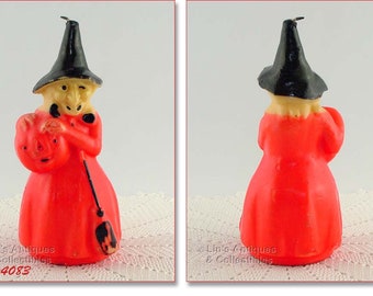 May include: A red and black Halloween witch candle with a black hat, holding a jack-o-lantern. The candle is on a white lace surface.