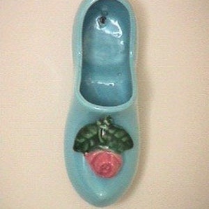May include: A light blue ceramic shoe planter with a pink rose and green leaves. The planter has a hole in the top for hanging.