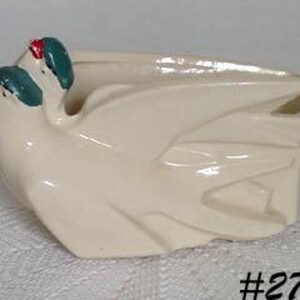 May include: Cream-colored ceramic planter with a stylized bird design. The planter features a geometric shape with a glossy finish. The bird's head is teal with red accents. The planter is likely for small plants or succulents.