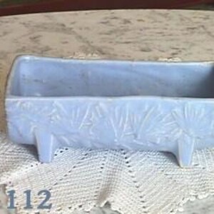 May include: A light blue rectangular planter with a textured floral design. The planter has four small legs and is sitting on a white lace tablecloth. The number "#1112" is visible in the image.