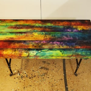 May include: A colorful wooden table with a rainbow-like paint design. The table has a rectangular top and black metal legs.