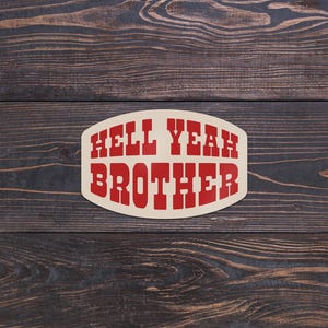 May include: An oval sticker with the words "HELL YEAH BROTHER" in red, bold, block letters. The sticker has a cream-colored background and is set against a dark wood grain surface.
