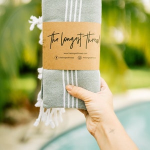 May include: A rolled up green and white striped Turkish towel with white fringe. The towel is wrapped in brown paper with the text "The Longest Thread" and the website address "www.thelongestthread.com".