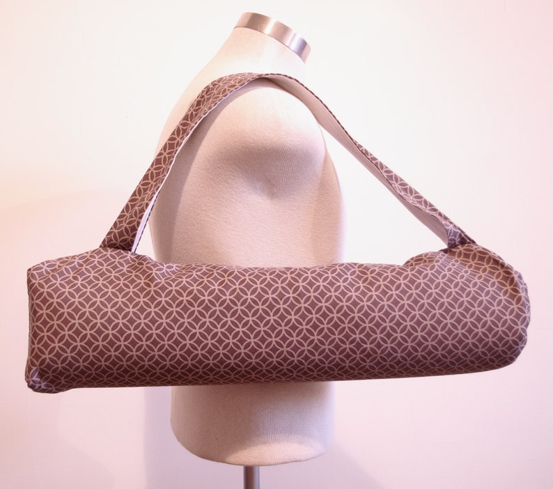 May include: A cylindrical yoga mat bag featuring a brown fabric exterior with a repeating white geometric circle pattern. The bag includes a long, integrated shoulder strap made from the same patterned fabric, designed for carrying a rolled mat.