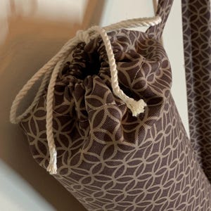 May include: A cylindrical fabric bag featuring a brown and beige geometric interlocking circle pattern. The bag includes a white twisted rope drawstring closure at the top and a matching fabric shoulder strap attached to the side of the bag.