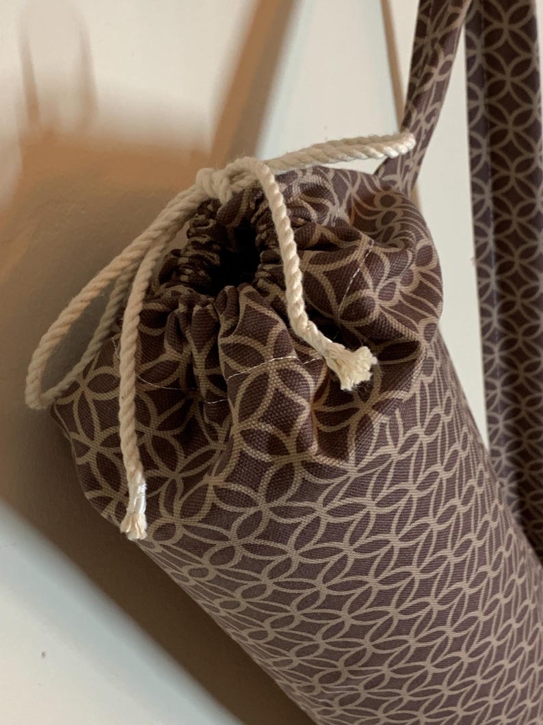 May include: A cylindrical fabric bag featuring a brown and beige geometric interlocking circle pattern. The bag includes a white twisted rope drawstring closure at the top and a matching fabric shoulder strap attached to the side of the bag.