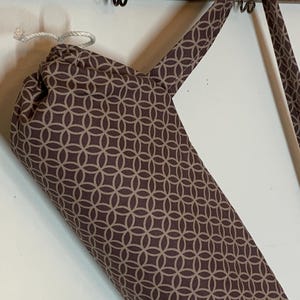 May include: A cylindrical fabric bag featuring a repeating brown and beige geometric circle pattern. The bag includes a white drawstring closure at the top and a matching fabric shoulder strap, hanging from a hook by its strap.