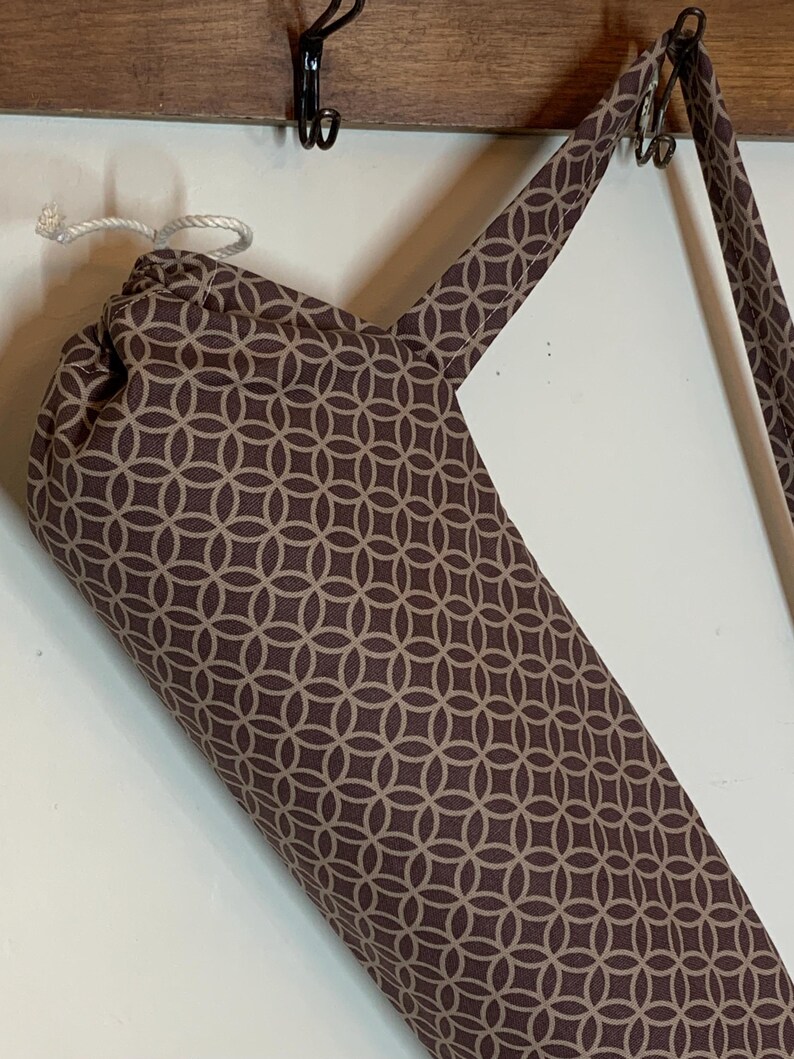 May include: A cylindrical fabric bag featuring a repeating brown and beige geometric circle pattern. The bag includes a white drawstring closure at the top and a matching fabric shoulder strap, hanging from a hook by its strap.