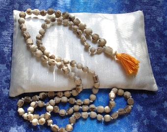 May include: A wooden mala bead necklace with a tassel on a white pillow. The beads are light brown and the tassel is orange.