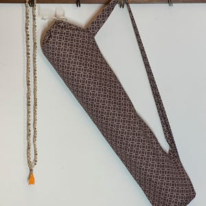 May include: A long, cylindrical fabric bag with a brown and white geometric circle pattern and a shoulder strap, hanging alongside a long string of small, light-colored wooden beads that features a single bright orange tassel at the bottom end.