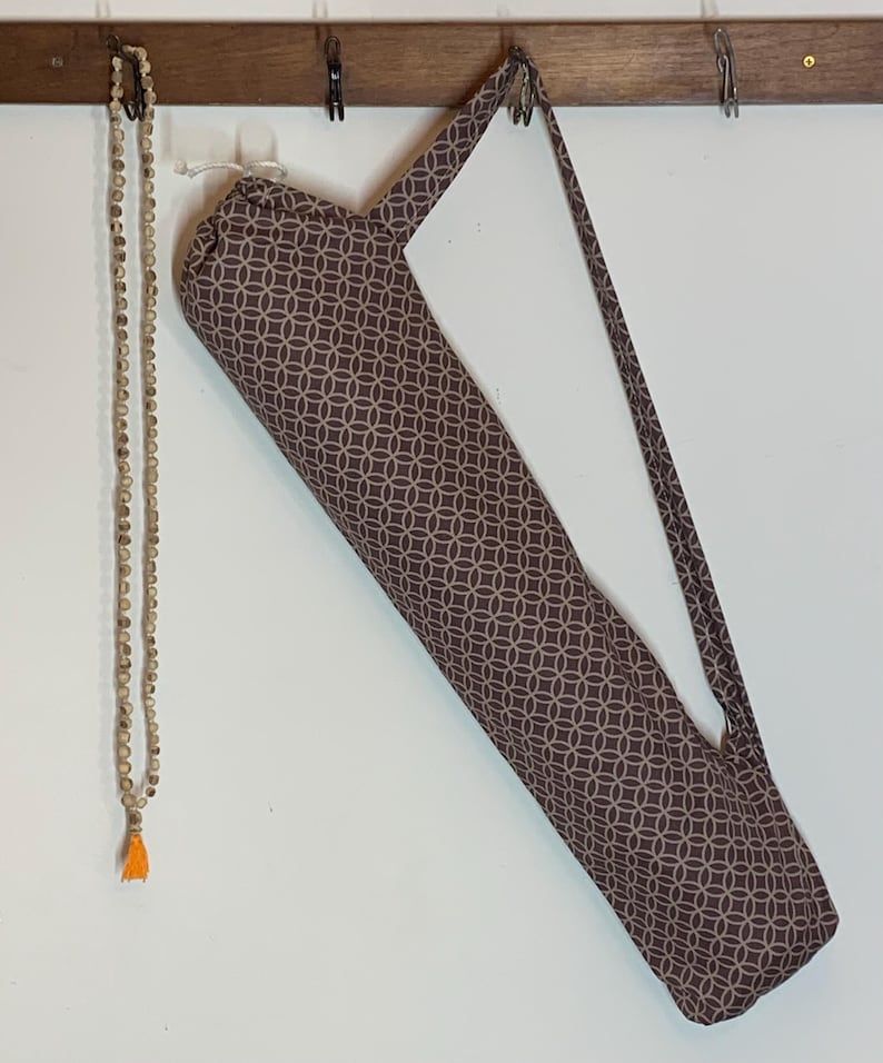 May include: A long, cylindrical fabric bag with a brown and white geometric circle pattern and a shoulder strap, hanging alongside a long string of small, light-colored wooden beads that features a single bright orange tassel at the bottom end.