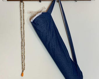 May include: A denim yoga mat bag with a white interior and strap hangs from a wooden rack. A beaded mala necklace with an orange tassel hangs to the left. The bag is a deep blue color.