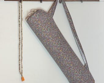 May include: A yoga mat bag with a colorful floral pattern, hanging on a wooden rack. A beaded necklace with an orange tassel hangs to the left. The bag has a shoulder strap and a zipper closure.