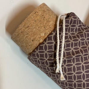 May include: A cylindrical container featuring a natural cork stopper and a brown fabric cover with a repeating beige geometric circle pattern. The fabric is secured at the top with two white twisted rope drawstrings hanging down the side.