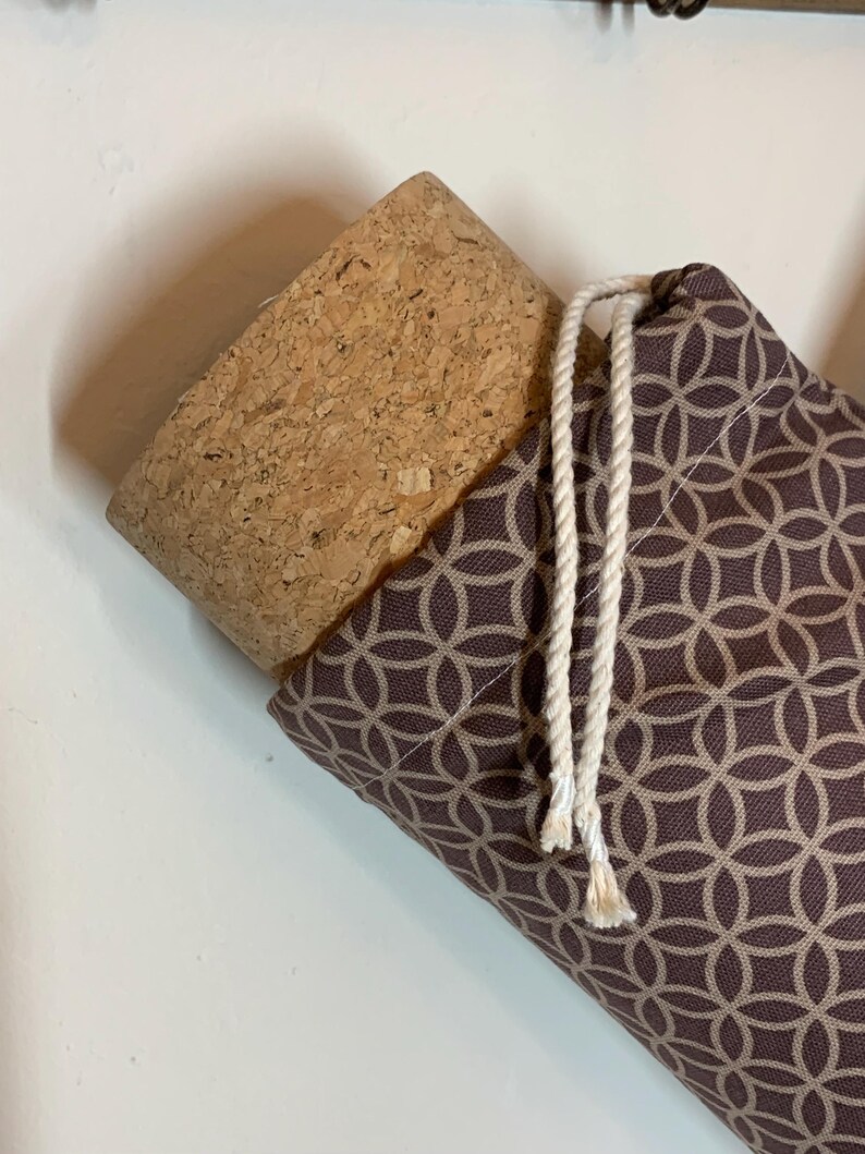 May include: A cylindrical container featuring a natural cork stopper and a brown fabric cover with a repeating beige geometric circle pattern. The fabric is secured at the top with two white twisted rope drawstrings hanging down the side.