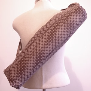 May include: A cylindrical yoga mat bag featuring a repeating geometric pattern of interlocking circles in light gray on a dark gray background. The bag has a long shoulder strap and is shown draped diagonally across a neutral-colored mannequin torso.