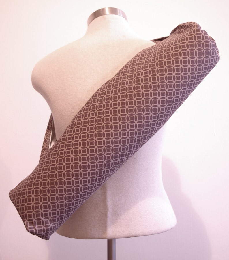May include: A cylindrical yoga mat bag featuring a repeating geometric pattern of interlocking circles in light gray on a dark gray background. The bag has a long shoulder strap and is shown draped diagonally across a neutral-colored mannequin torso.