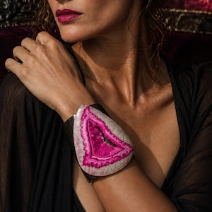 May include: A large, statement bracelet featuring a polished, pink and white agate stone set in a dark metal cuff. The stone has a striking, crystalline center and a unique, organic shape. The bracelet is worn on the wrist.