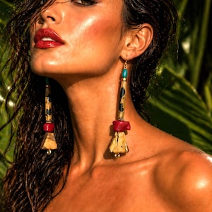 May include: Long, dangling earrings with a unique design. The earrings feature a combination of materials, including wood, red and turquoise beads, and gold-colored metal accents. The earrings have a tribal or bohemian aesthetic.