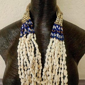 May include: A handcrafted necklace featuring strands of white seashells cascading from a woven beige base. The necklace incorporates blue and white beaded accents, creating a striking contrast. The necklace is displayed on a dark mannequin.