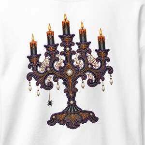 May include: A white long-sleeve sweatshirt featuring a central graphic of an ornate, dark purple and gold candelabra with five lit black candles. The design includes intricate spiderweb patterns, small hanging white beads, and a dangling black spider.