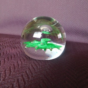 Puede incluir: Un pisapapeles de vidrio transparente con una burbuja verde en su interior. La burbuja tiene una forma abstracta y en espiral.