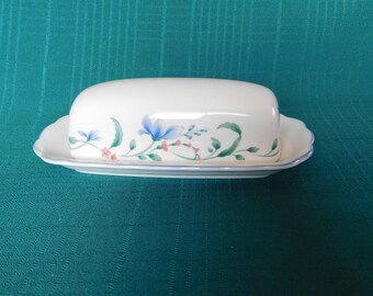 Könnte beinhalten: Eine weiße Keramik-Butterdose mit einem Blumendesign in Blautönen, Grün und Rosa. Die Dose hat einen Deckel und einen gewellten Rand.