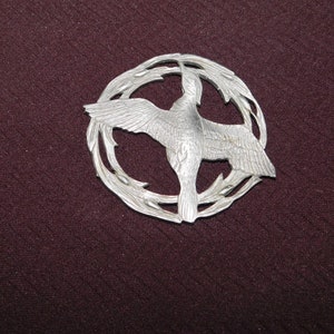 May include: A silver-toned metal pin featuring a stylised bird in flight, surrounded by a circular design of stylised branches.