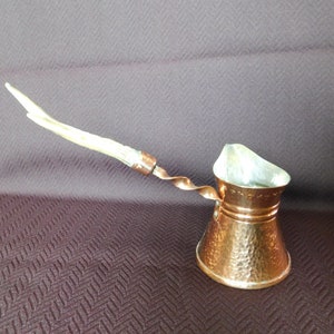 May include: A copper coffee pot with a long, curved handle. The pot has a hammered texture and a twisted handle attachment. The handle is light-coloured, possibly bone or wood. The pot is designed for brewing coffee.