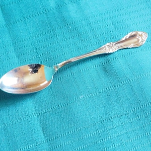 Può includere: Un cucchiaino d'argento con un manico decorato. Il cucchiaino è appoggiato su una superficie di tessuto blu turchese.