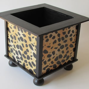 May include: A black wooden planter with a leopard print design. The planter has four rounded legs and a square opening at the top.
