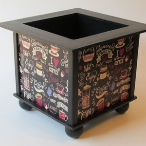 May include: Black wooden box with a coffee themed design. The box has a square shape and four rounded legs. The design features coffee cups, coffee beans, and text that says "I love coffee" and "Hot coffee".