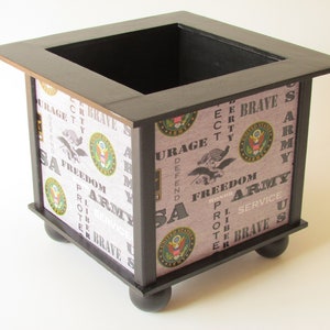 May include: A square wooden box with a black frame and feet. The sides are covered in a patterned fabric featuring the words "Army", "Freedom", and "Courage", along with the US Army emblem. The box has a dark interior.