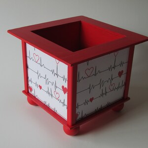 May include: Red wooden box with a white background and a pattern of black heartbeats and red hearts. The box has four small feet.