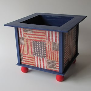 May include: A blue wooden planter box with red feet. The box is decorated with a patchwork of American flag designs.