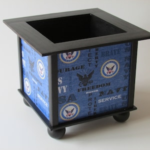 May include: A black wooden planter with a blue and white patterned liner featuring the US Navy seal and words like "Freedom", "Honor", "Service", and "Brave".