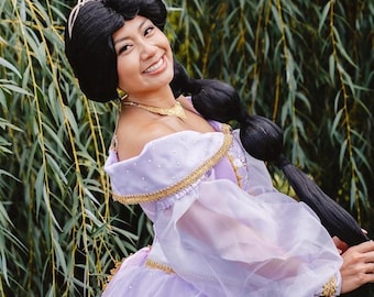 May include: A lavender off-the-shoulder gown featuring sheer puff sleeves, gold lace trim, and scattered pearl accents. The ensemble includes a gold necklace, gold earrings, and a gold tiara worn with a long black wig styled in a low ponytail.