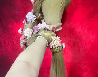 May include: A long, golden blonde wig with a braid and floral accents. The wig is styled with a long braid and adorned with pink and purple flowers. The wig is on a mannequin head against a red background.