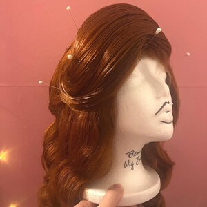 May include: A long, wavy, copper-red wig styled with a side-swept fringe and secured with several white-headed straight pins. The wig is displayed on a white foam mannequin head with the text "Blonde Wig Bar" printed on the neck area.