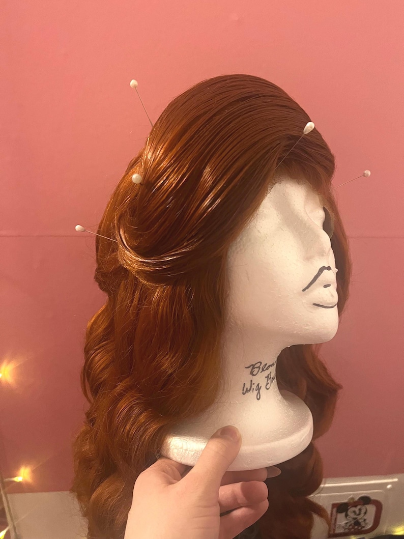 May include: A long, wavy, copper-red wig styled with a side-swept fringe and secured with several white-headed straight pins. The wig is displayed on a white foam mannequin head with the text "Blonde Wig Bar" printed on the neck area.
