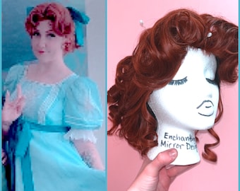 May include: A red wig with curls styled on a mannequin head. The wig is on a pink background. The mannequin head has the words "Enchanting Mirror Designs" written on it.