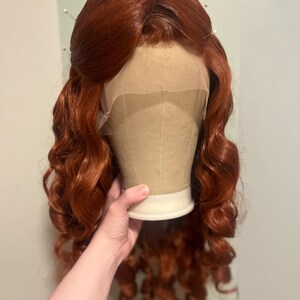 May include: A long, wavy, copper-red wig styled with a centre part and soft curls cascading down the sides. The wig features a sheer lace front cap construction, visible at the hairline, and is secured with several straight pins at the crown.