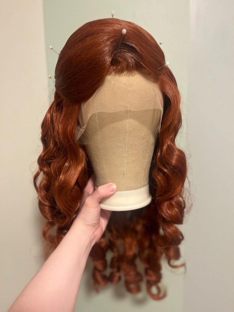 May include: A long, wavy, copper-red wig styled with a centre part and soft curls cascading down the sides. The wig features a sheer lace front cap construction, visible at the hairline, and is secured with several straight pins at the crown.