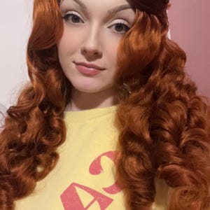 May include: A long, vibrant copper-red wig featuring voluminous, tight curls that cascade down the shoulders. The hair is styled with a smooth, swept-back crown section. The wig is worn over a pale yellow shirt with pink text printed on the front.