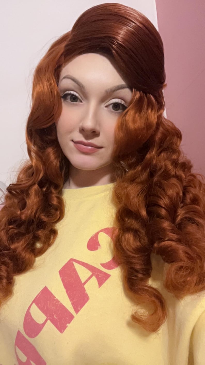 May include: A long, vibrant copper-red wig featuring voluminous, tight curls that cascade down the shoulders. The hair is styled with a smooth, swept-back crown section. The wig is worn over a pale yellow shirt with pink text printed on the front.