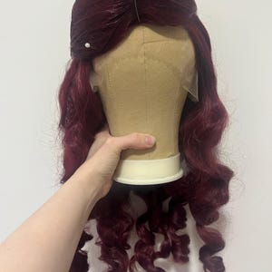 May include: A deep burgundy wig with long, loose curls styled on a beige mannequin head. The wig features a lace front hairline and is secured at the top with three white-headed sewing pins, showcasing a voluminous, wavy texture throughout.