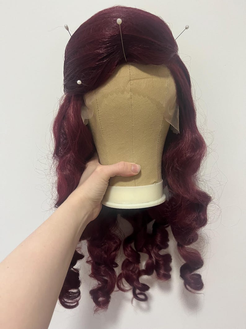 May include: A deep burgundy wig with long, loose curls styled on a beige mannequin head. The wig features a lace front hairline and is secured at the top with three white-headed sewing pins, showcasing a voluminous, wavy texture throughout.